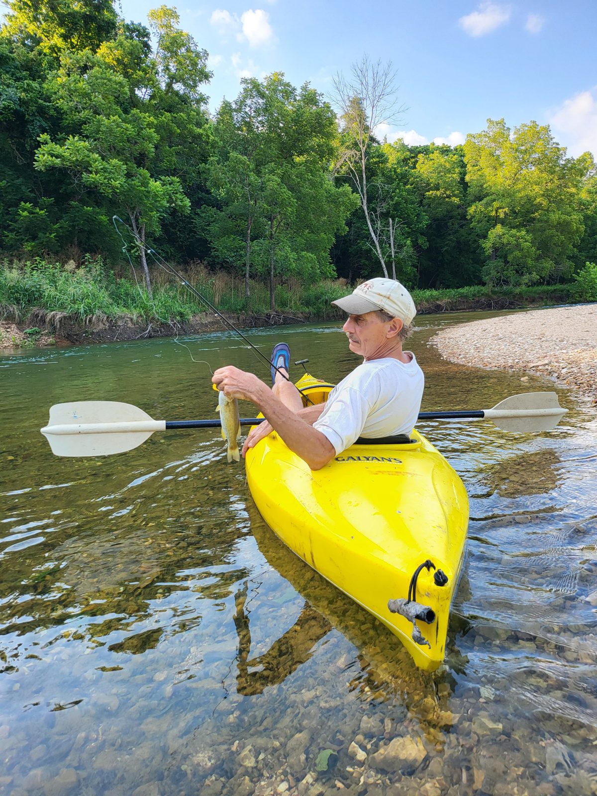 A kayaker holding a freshly caught fish beside a yellow kayak on the clear water of Bull Creek